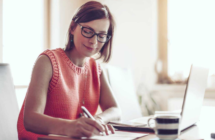 Woman sitting in front of computer smiling and writing on paper
